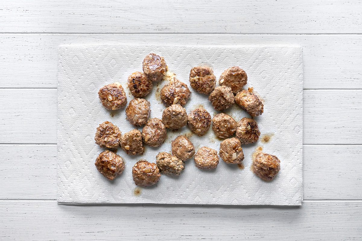 overhead shot of Several cooked meatballs are arranged on a white paper towel, which is placed on a white wooden surface, The meatballs appear browned and slightly oily