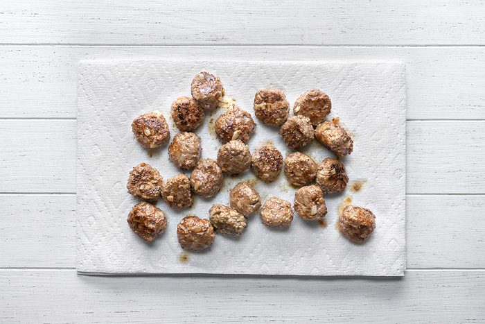 overhead shot of Several cooked meatballs are arranged on a white paper towel, which is placed on a white wooden surface, The meatballs appear browned and slightly oily