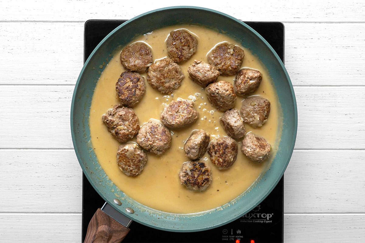 overhead shot of Meatballs simmering in a creamy brown sauce in a blue skillet on an electric stovetop, viewed from above, on a white wooden surface