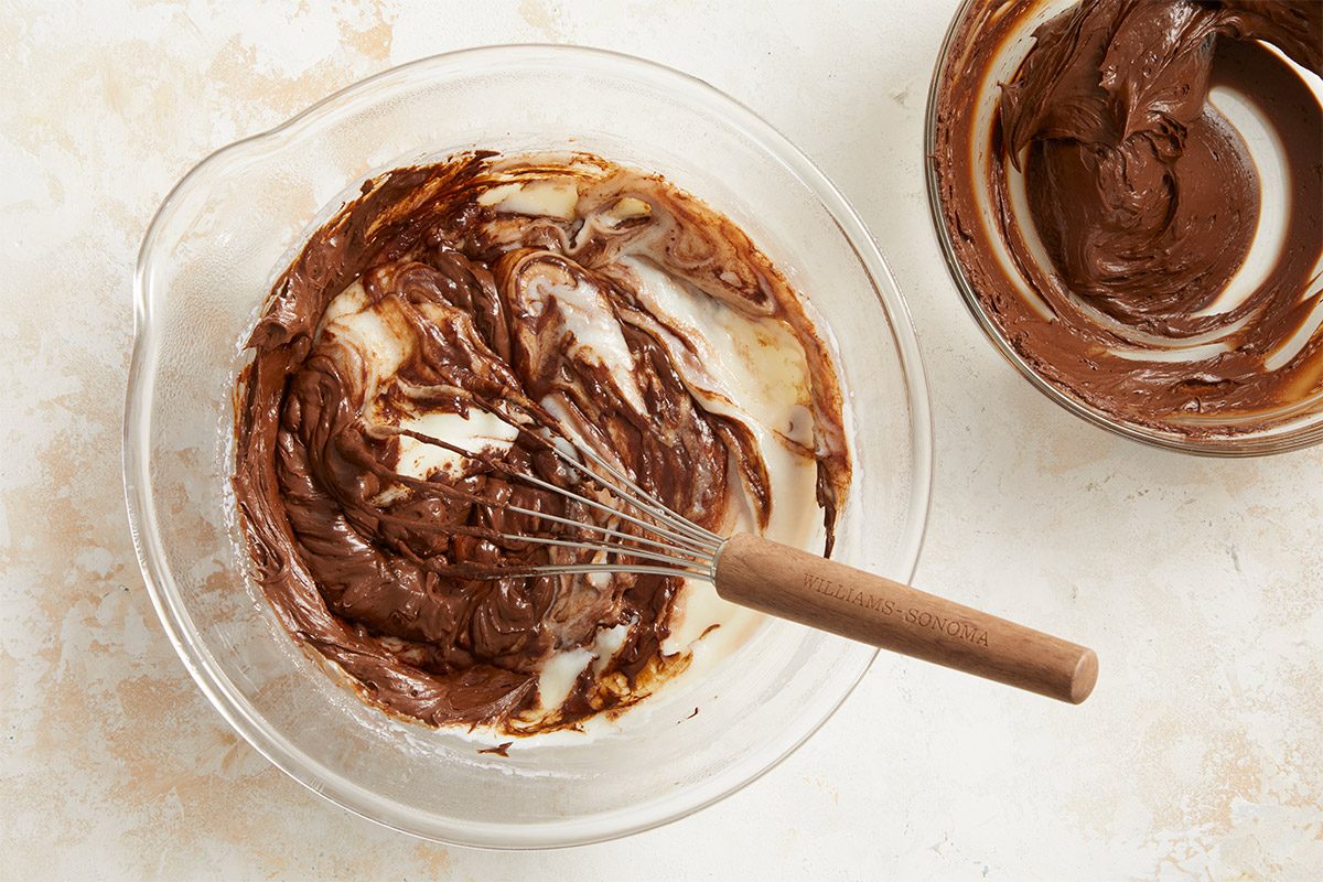 Overhead shot of chocolate brownie batter and milk being whisked together in a glass mixing bowl, with a second bowl of batter nearby;