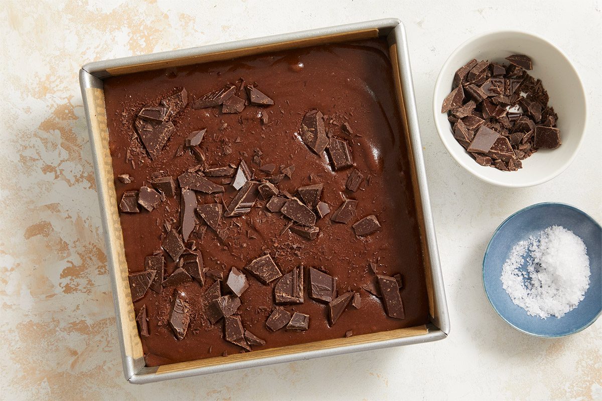 Overhead shot of chocolate brownie batter in a square baking pan topped with chopped chocolate, with bowls of extra chocolate and flaky salt on the side;