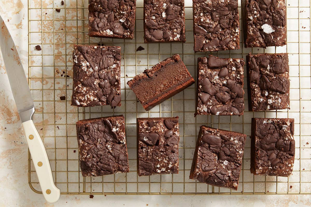Overhead shot of sliced mochi brownies arranged on a cooling rack, with one brownie turned on its side to reveal its chewy interior; a knife rests beside the rack;