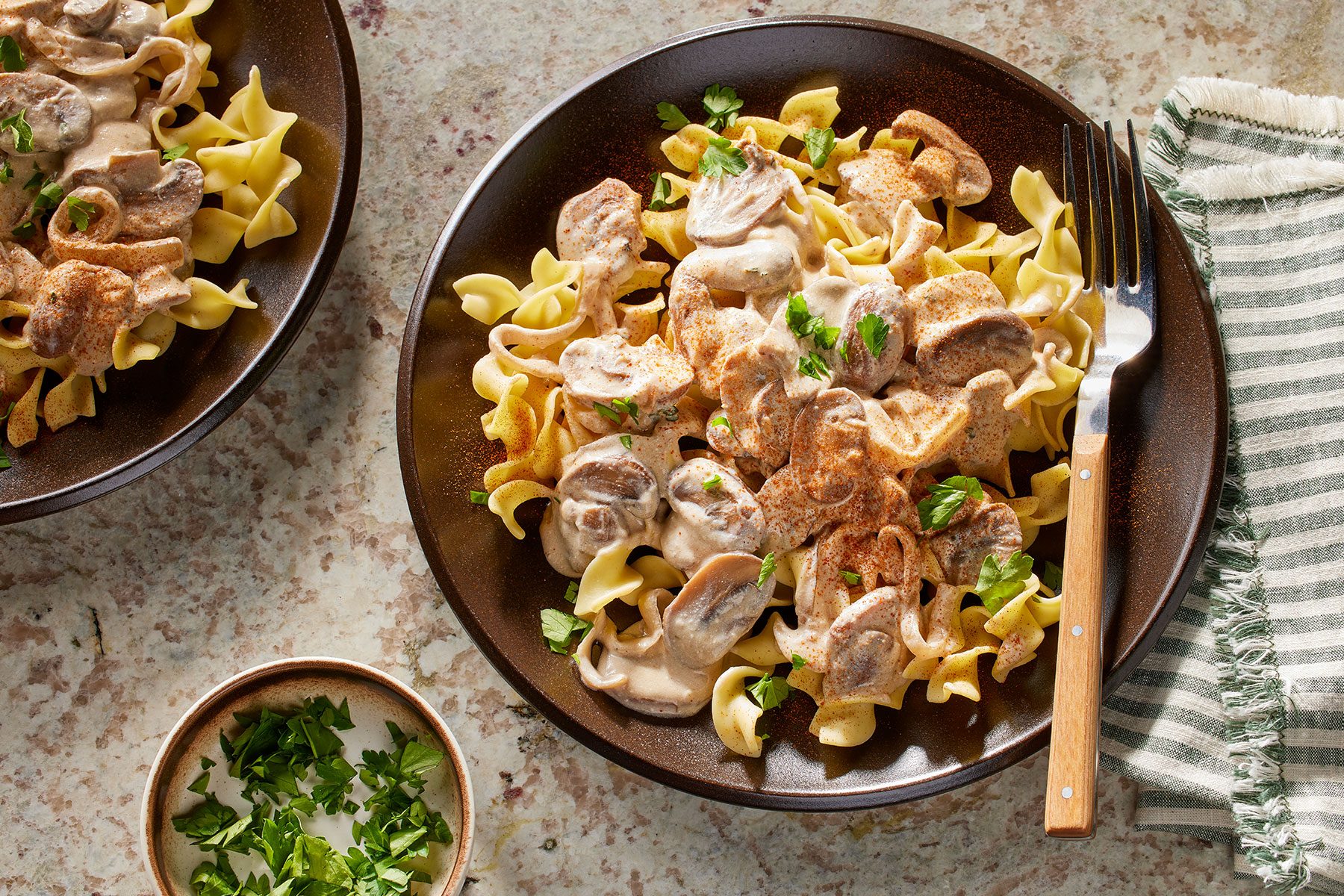 Mushroom Stroganoff served in small plates with a fork and some garnishing in a bowl next to it.
