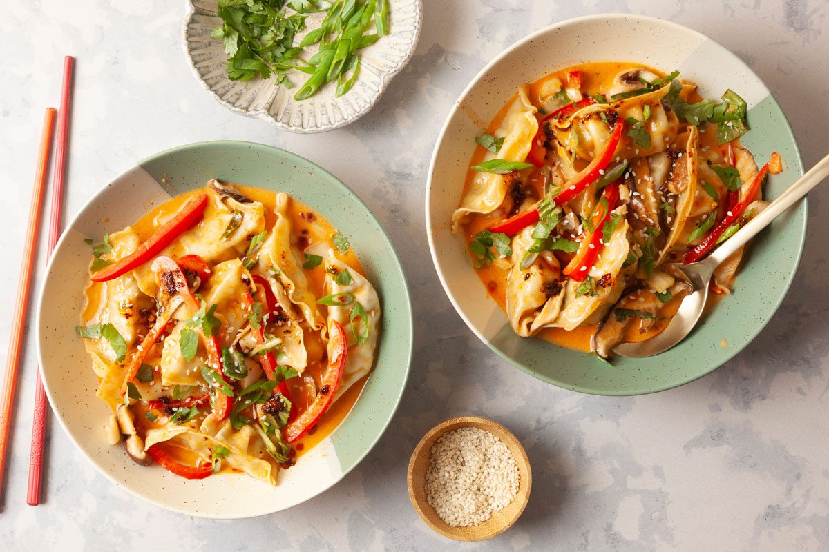 Overhead shot of Two bowls of One Pan Dumpling Dinner with vegetables basil and sesame seeds sit near sesame seeds green onions and red chopsticks