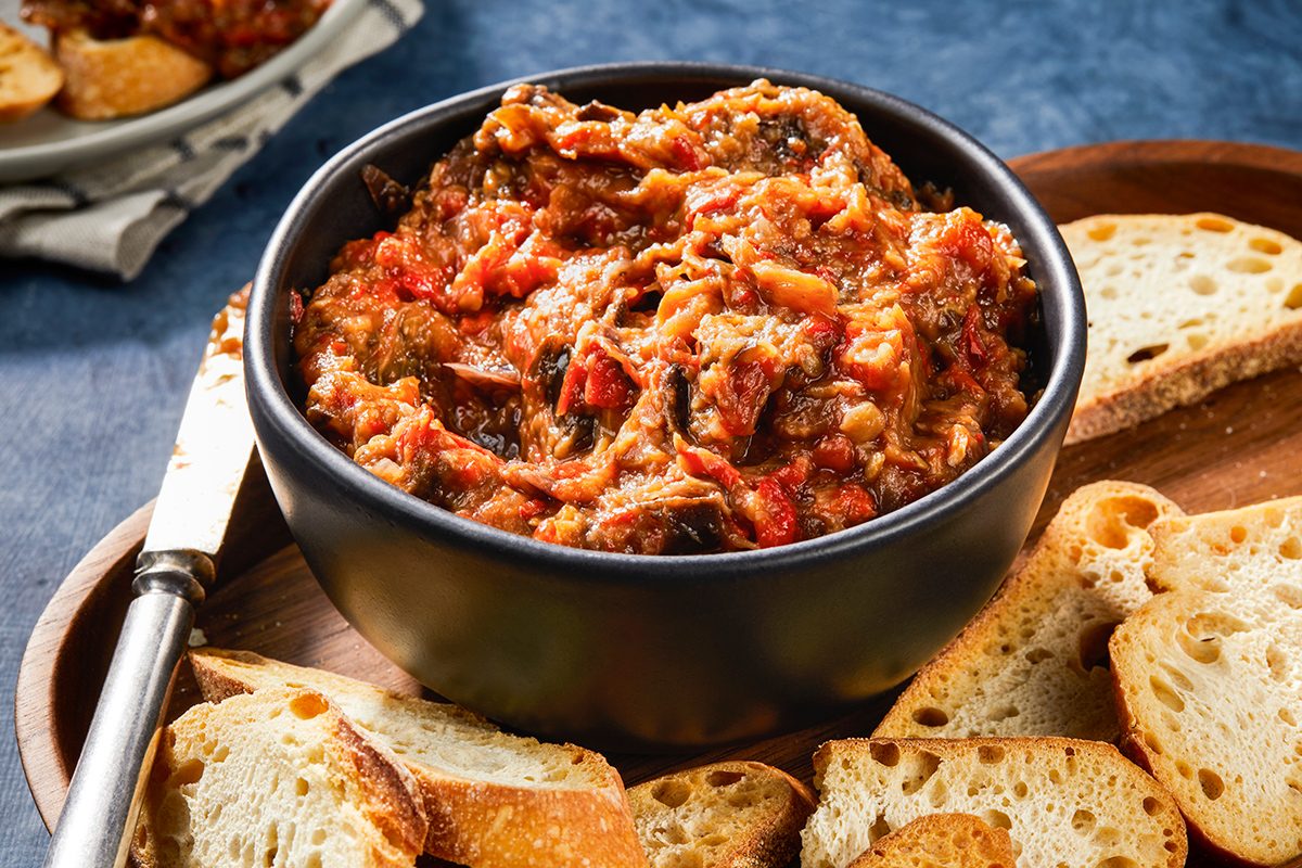 A black bowl filled with chunky vegetable and tomato dip is surrounded by slices of toasted bread on a wooden tray, with a knife resting nearby.