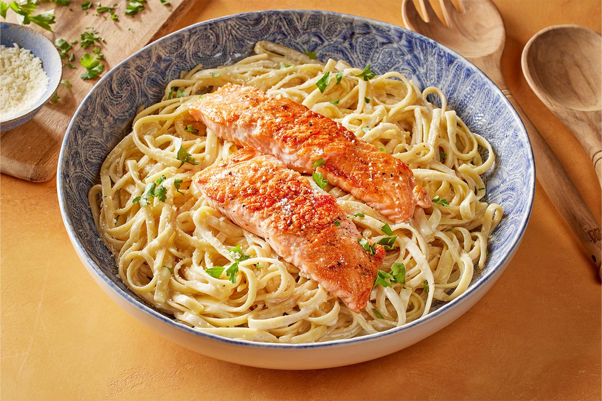 Close shot of a Salmon Alfredo featuring a bowl of creamy fettuccine with two grilled salmon fillets topped with green parsley nearby are wooden utensils and a bowl of grated cheese the surface below is light colored and smooth giving the scene a bright clean feel