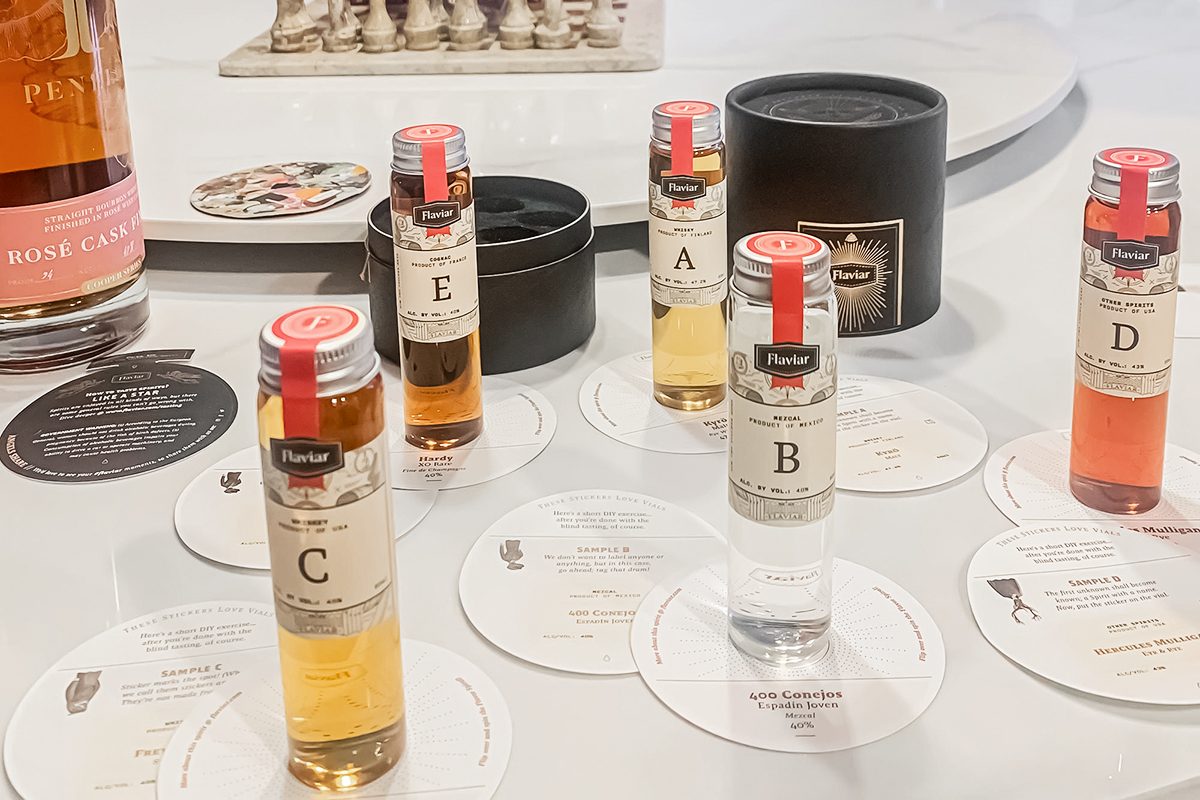 Five labeled sample vials of whiskey stand on circular tasting cards, with a round black container and a bottle of rosé in the background, all arranged on a white surface.