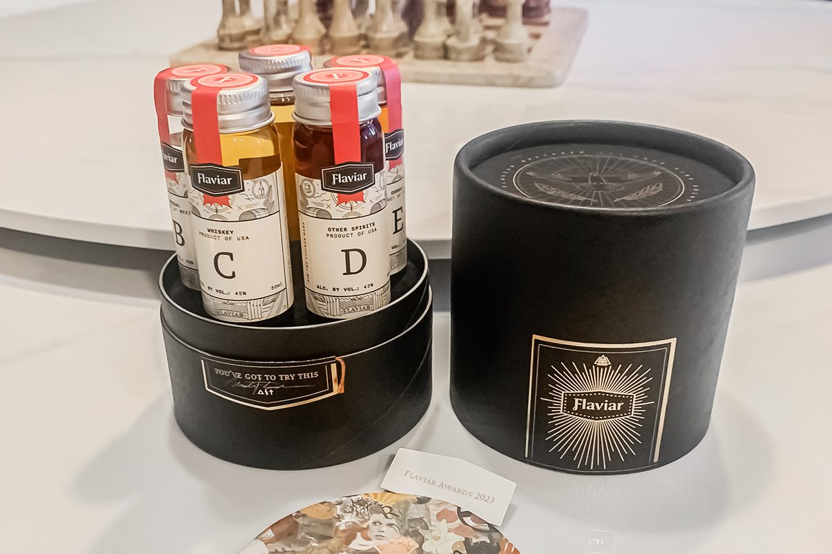 A round black gift box labeled "Flaviar" holds five small glass vials of spirits, each with a letter label. The box lid and a coaster are placed beside it on a light-colored surface.