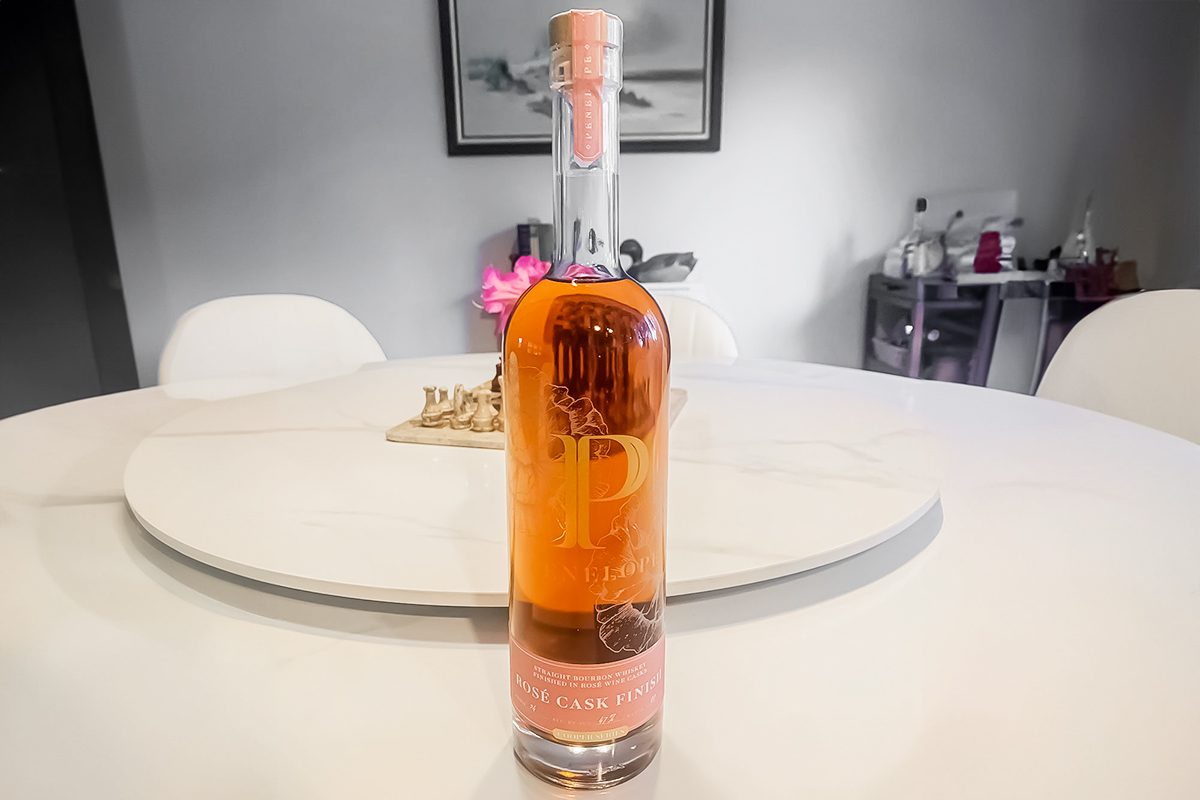 A bottle of rosé wine with a pink label and cap sits on a white round table in a modern dining room with white chairs and a blurred background.