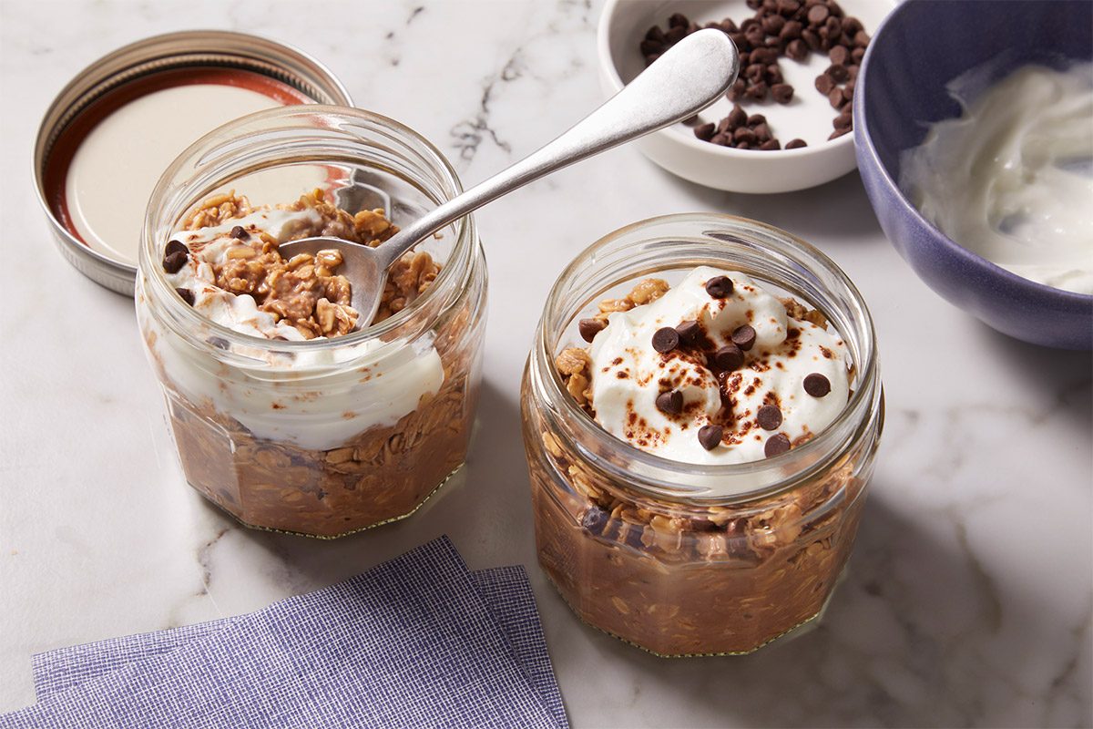 Close overhead shot of two jars of Tiramisu Overnight Oats with a spoon scooping into the creamy oat mixture, showing the thick texture and cocoa-dusted topping.