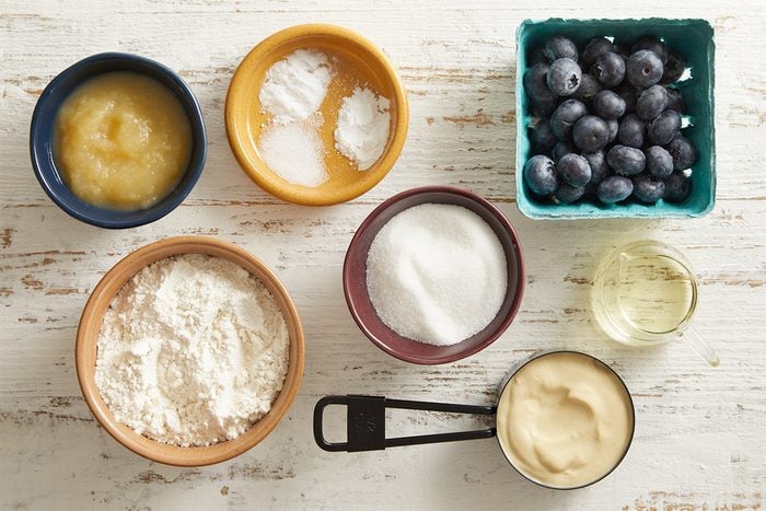 Assorted baking ingredients in bowls and cups on a white wooden surface, including blueberries, flour, sugar, applesauce, baking powder, baking soda, salt, oil, and a creamy substance.