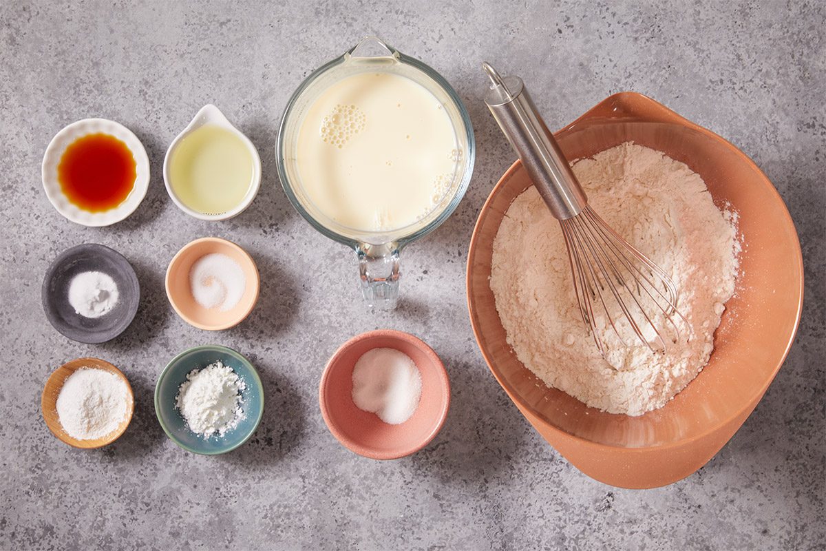 Overhead shot of baking ingredients are spread out on a gray countertop A bowl of flour with a whisk sits beside milk and small bowls of vanilla oil sugar salt baking powder and cornstarch